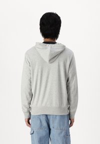Hellgrauer Kapuzenpullover aus weichem Material, mit Kapuze, lockerem Schnitt und geripptem Saum. Kombiniert mit blauen weiten Jeans.