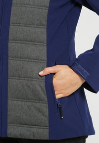 Marinblå softshell-jacka med en grå vadderad panel, zippered ficka och velcro-muddar. Slät textur med en figursydd design.