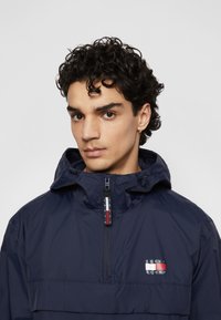 Navy windbreaker with a half-zip design, adjustable hood, and logo patch. Features smooth texture and a contrasting zipper pull.