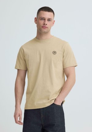 Uomo con capelli corti che indossa una maglietta beige a maniche corte e jeans scuri, in piedi con una mano in tasca contro uno sfondo semplice.