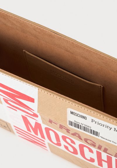 Sac beige rectangulaire ouvert ressemblant à une boîte en carton avec l'inscription rouge "FRAGILE" et "MOSCHINO" et une petite poche intérieure.