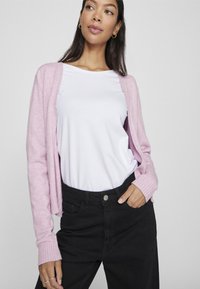 Cardigan rosa chiaro con polsini a coste, indossato aperto su una t-shirt bianca e pantaloni neri a vita alta. Tessuto morbido, vestibilità casual.
