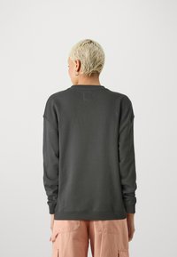 Sudadera gris oscuro con mangas largas, puños acanalados y cuello redondo. Material suave con una textura sutil y un pequeño parche con logo en la espalda.