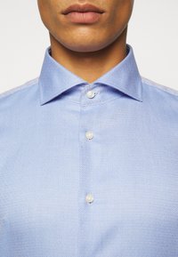 Camicia a maniche lunghe di colore blu chiaro con motivo a diamante, colletto puntato e bottoni bianchi. Primo piano del colletto e della parte anteriore.