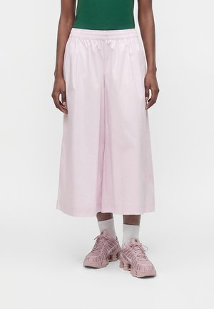 Personne portant un pantalon large rose pâle coupé, des chaussettes blanches et des baskets roses, debout contre un fond blanc.