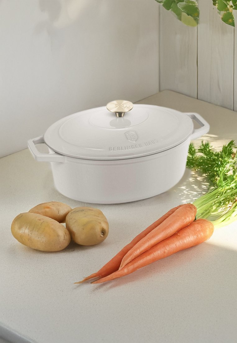 Weißer keramischer Dutch Oven mit runder Form, goldener Knauf und eingeprägtem Markenlogo, neben drei Kartoffeln und zwei orangen Karotten auf einer Arbeitsplatte.