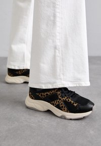 Baskets à imprimé léopard noir et fauve en matériaux mesh et cuir, disposant d'une semelle crème coussinée et de lacets, associées à un pantalon blanc à jambes larges.