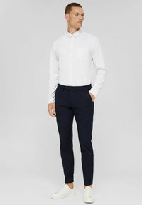 White long-sleeve button-up shirt with a pocket, paired with dark navy slim-fit trousers and white sneakers. Smooth fabric, tailored design.