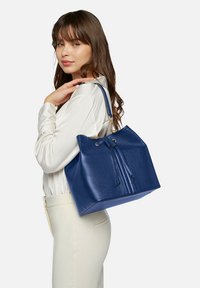 Borsa a secchiello in pelle blu con chiusura a coulisse, superficie testurizzata e dettagli in metallo dorato. Spallacci robusti per il trasporto.