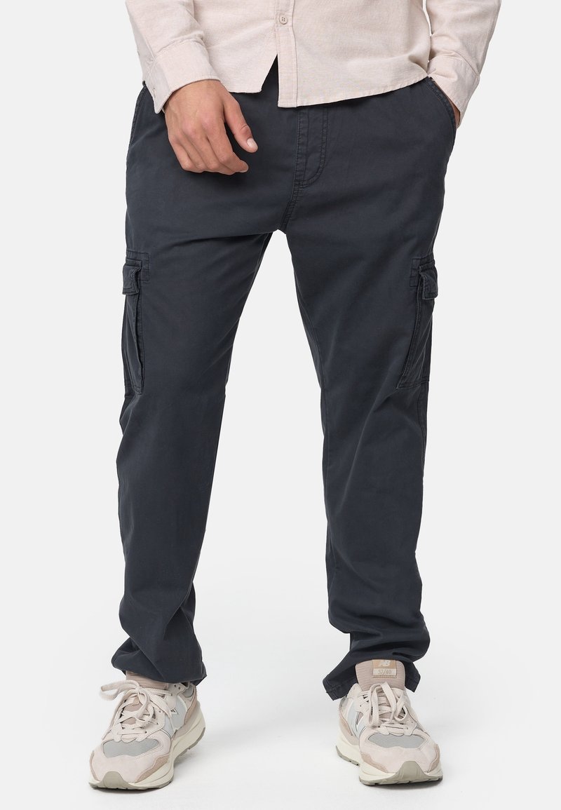 Homme portant un pantalon cargo bleu marine foncé avec des poches latérales, une chemise beige clair et des baskets beiges, debout devant un fond blanc.