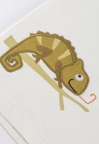 Weißer Stoff mit einer detaillierten Stickerei eines Chamäleons in Grün- und Brauntönen, mit einer oranger Zunge, die auf einem Ast sitzt.