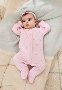 Rožinė kūdikių kombinezonas su mažais raudonais širdelių raštais, baltu apykaklėle ir priekiniais mygtukais. Minkšta medžiaga, uždara avalynė ir ilgomis rankovėmis.