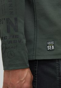 Camicia a maniche lunghe in cotone verde scuro con testo stampato, polsini elastici e un'etichetta con "Camp David Arctic Sea" sull'orlo.