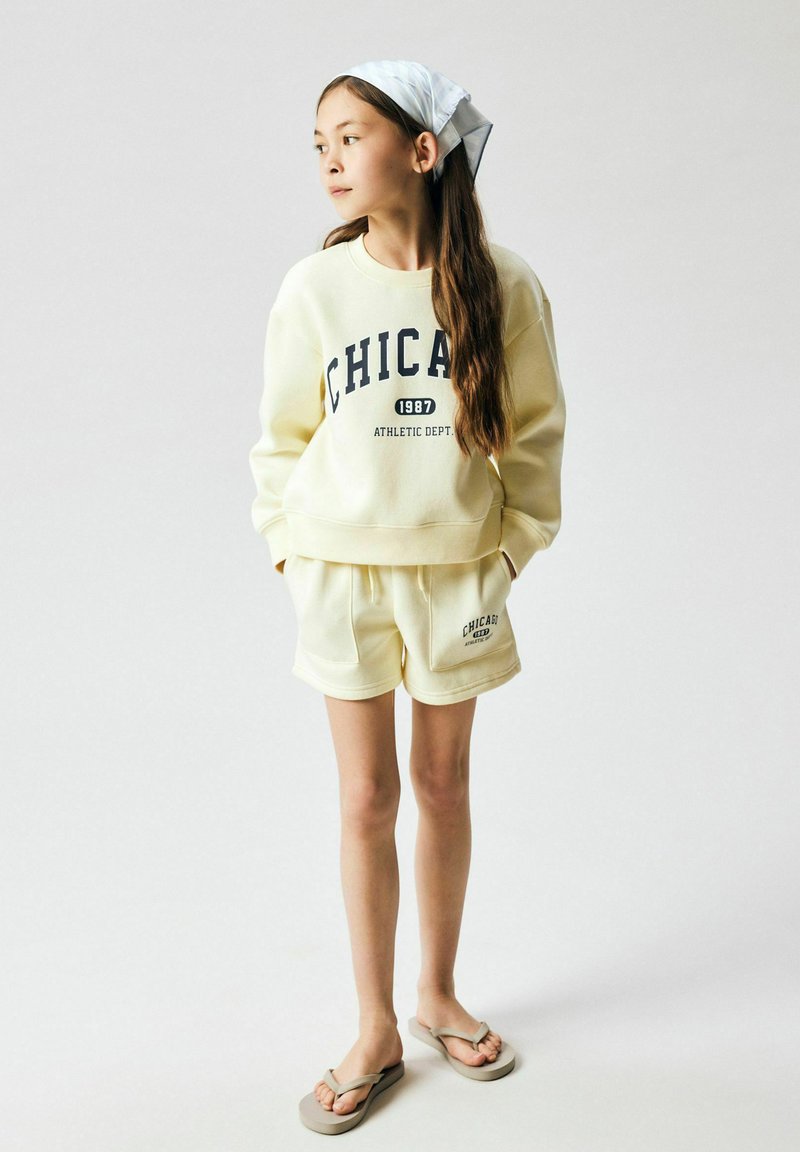 Jeune fille debout regardant vers la gauche, portant un sweat-shirt crème « CHICAGO » et un short assorti, un foulard gris sur la tête et des tongs beige sur fond blanc.