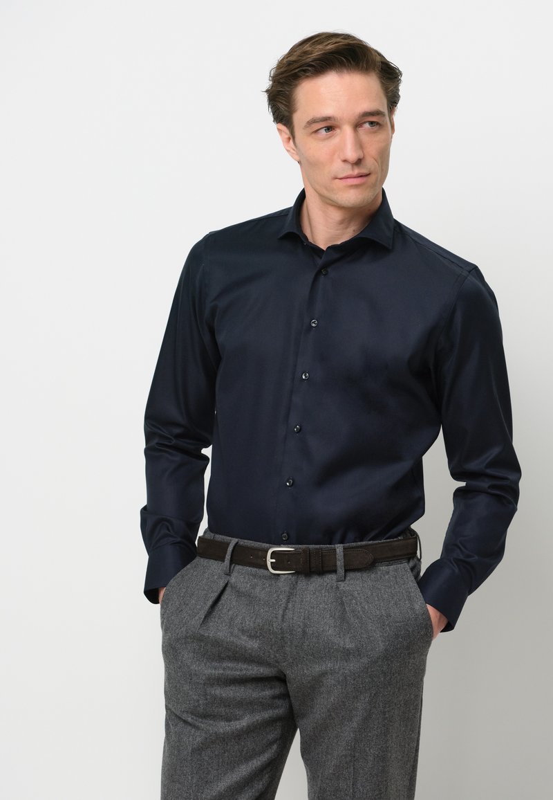 Marineblauw, langemouwenshirt met een knoopsluiting aan de voorkant, gecombineerd met lichtgrijze, gestructureerde broeken en een bruine riem met een zilveren gesp.