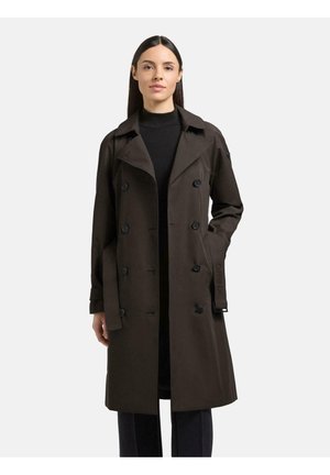 Frau mit langen dunklen Haaren, die einen dunkelbraunen, doppelreihigen Trenchcoat über schwarzer Kleidung trägt und mit einer Hand in der Tasche steht.