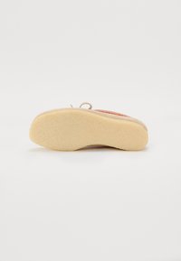 Chaussure avec une semelle beige, texturée et épaisse pour une bonne adhérence. Le dessus a des accents tissés colorés et un design arrondi avec un détail en boucle centrale.