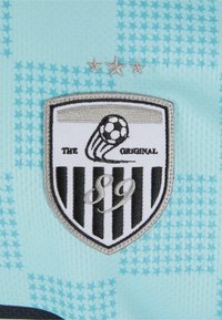 Izšūts uzlīmes dizains ir šahtas forma ar melnbaltu svītrojumu, futbolbumbas grafiku un tekstu "ORĢINĀLS" virs "89", uz gaiši zila fona.