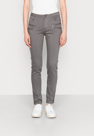 Pantalones grises ajustados con cuatro bolsillos delanteros con cremallera y cremalleras en los tobillos, usados con zapatillas blancas y una camiseta blanca.