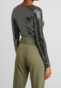 Schwarzes Paillettenoberteil mit langen Ärmeln und Chevron-Muster, kombiniert mit olivgrünen, hochgeschnittenen Hosen mit glatter Textur und einer hinteren Tasche.