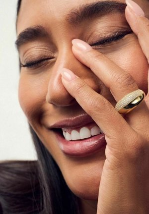 Guldring med en struktureret overflade, der har små indlagte sten. Hånden hviler på ansigtet og viser glat hud og naturlige negle.