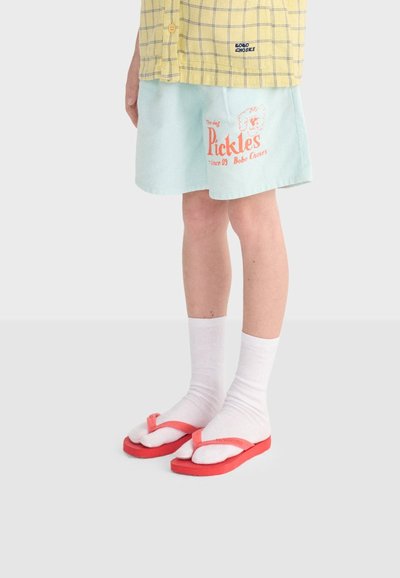 Enfant portant un short bleu clair avec texte et graphisme rouges, des chaussettes blanches longues, des tongs rouges et une chemise à carreaux jaune.