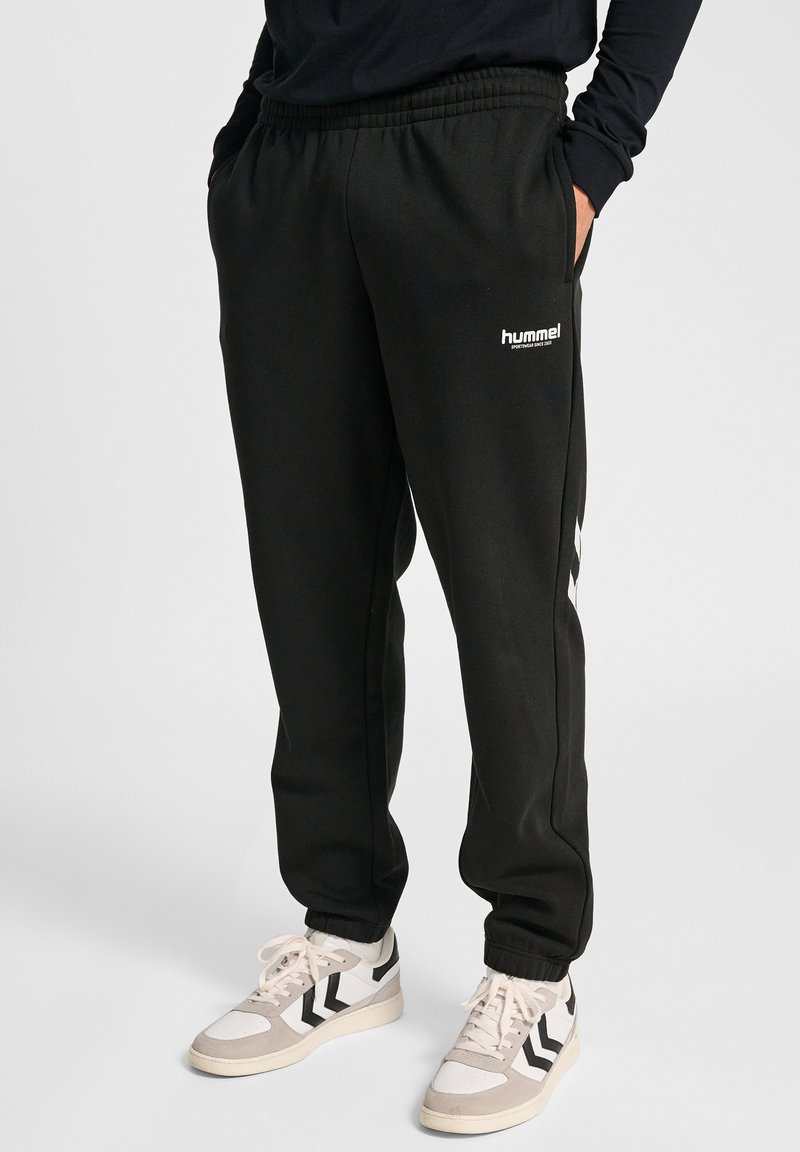 Zwarte sweatpants van een zachte stof, met een elastische tailleband, zijzakken en witte logo-details. Gecombineerd met lichtgekleurde sportschoenen.