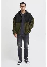 Unge mand iført en grøn og sort fleecejakke, mørkegrå jeans, sorte sneakers, stående mod en hvid baggrund med et neutralt udtryk.