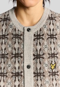 Cardigan lavorato a maglia di colore grigio chiaro con motivi marroni e neri, dotato di un colletto a costine e di un bottone nero all'apertura del collo. Accento con logo giallo.