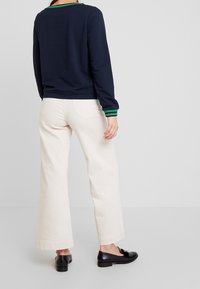 Námořnická mikina s manžetami s zeleno-černými pruhy, kombinovaná s krémovými širokými kalhotami a černými mokasíny, zobrazená zezadu.