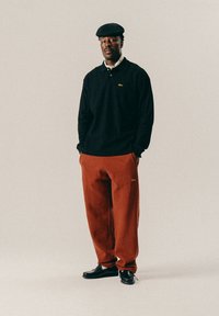 Man met een zwart languit shirt, roestkleurige broek, zwarte schoenen en een zwarte pet met platte kroon, staande met zijn handen in zijn zakken.