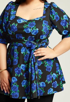 Donna che indossa un vestito nero con stampa floreale blu e maniche a sbuffo, legato in vita, con la mano appoggiata sul fianco e un piccolo tatuaggio nella parte superiore del petto.