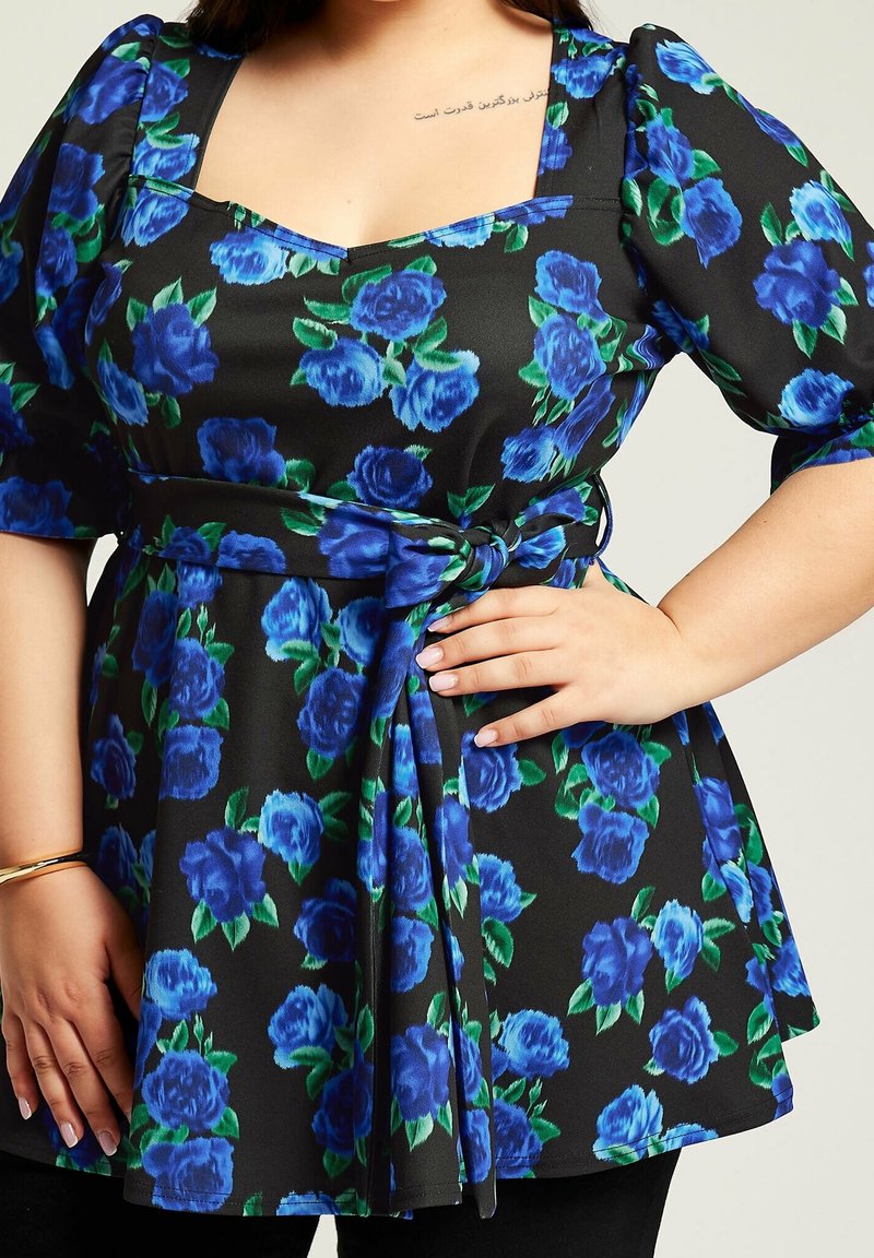 Femme portant une robe noire à imprimé floral bleu avec des manches bouffantes, nouée à la taille, la main posée sur la hanche et un petit tatouage sur le haut de la poitrine.