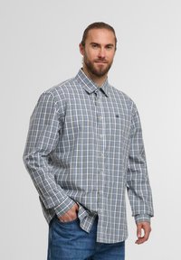 Mann mit Bart, der ein blau-weiß kariertes Langarmhemd mit Knöpfen und eine blaue Jeans trägt, und mit einer Hand in der Tasche steht.