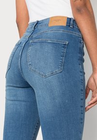 Jeans azuis de cintura alta com um efeito desbotado, apresentando um patch de couro castanho na parte de trás da cintura e dois bolsos traseiros.