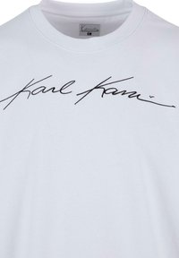 T-shirt en coton blanc avec un grand motif en noir de type signature en cursive au centre. Encolure ras du cou classique, texture douce.