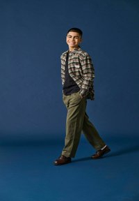 Geruite flanellen shirt in beige, groen en rood, zwart T-shirt, olijfgroene broek en bruine schoenen, staand tegen een blauwe achtergrond.