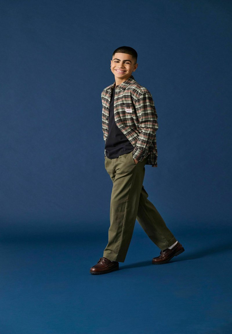 Geruite flanellen shirt in beige, groen en rood, zwart T-shirt, olijfgroene broek en bruine schoenen, staand tegen een blauwe achtergrond.