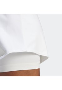 Top atlético blanco con dobladillo curvado, superpuesto a unos pantalones cortos ajustados blancos. Tela suave con un ligero brillo. Detalles de costura limpios.
