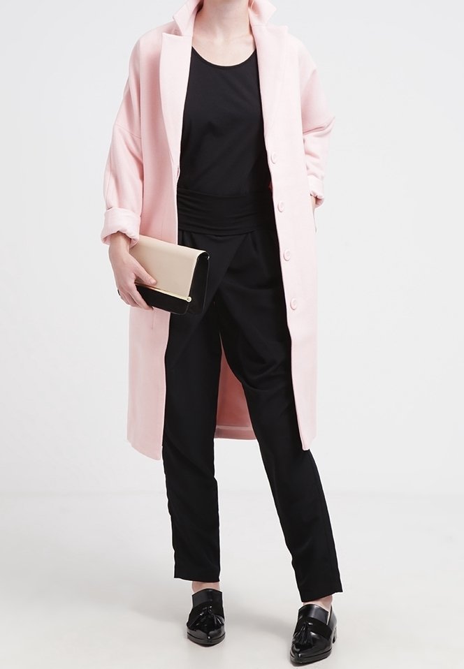 Long manteau rose à coupe décontractée, haut noir, pantalon noir ample, et mocassins noirs brillants, tenant une pochette beige.