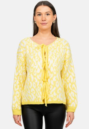 Woman wearing a yellow and white leopard-print cardigan with front tie closures and black leather pants, facing forward on a white background.