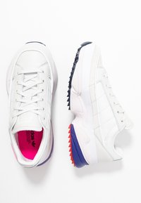 Vitaidiga skor i vitt med mesh- och syntetmaterial, en kontrasterande marinblå häl och en sula med mönster i rött och lila.
