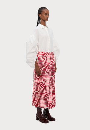 Chemise blanche à boutons avec des manches volumineuses, associée à une jupe rouge et blanche à motifs avec des rayures ondulées ; bottes à talons noires brillantes.