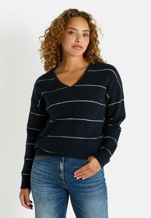 Suéter de punto azul marino con franjas horizontales doradas, diseño de escote en V, ajuste relajado y puños acanalados. Combinado con jeans de mezclilla azul claro.