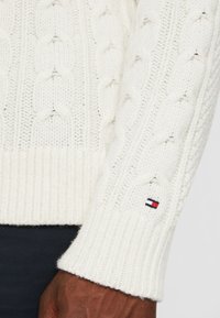 Cremefarbener Kabelstrickpullover mit gerippten Bündchen und Saum. Kleines gesticktes Logo in Rot, Weiß und Blau nahe dem Bündchen. Weicher, strukturierter Stoff.