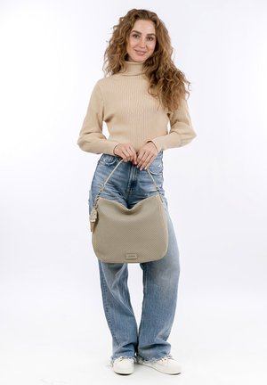 Bolso de mano de cuero perforado beige con una silueta curva y una correa corta, que presenta un pequeño detalle de etiqueta. El modelo lleva puestos unos jeans de color azul claro y un suéter beige.