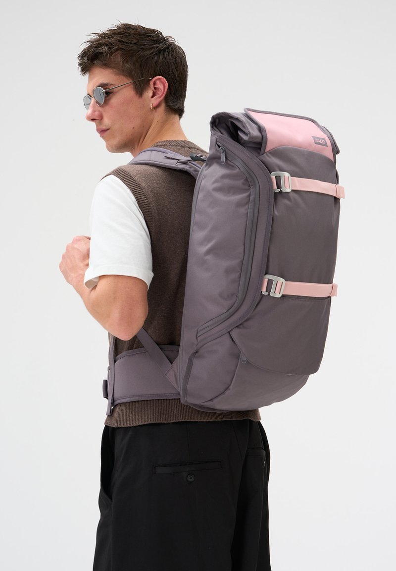 Un homme portant des lunettes de soleil et un gilet marron porte un grand sac à dos de voyage gris et rose avec des bretelles rembourrées et une ceinture ventrale.