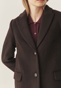 Femme portant un manteau en laine brun foncé sur une chemise bordeaux boutonnée, debout avec un bras derrière le dos, sur un fond neutre.