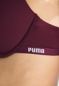 Burgundski sportski grudnjak od glatkog materijala s PUMA logotipom u bijeloj boji na teksturiranoj elastičnoj traci, s okruglim dekolteom i ojačanim šavovima.