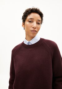 Bordeauxroter Pullover mit geripptem Rundhalsausschnitt und Raglanärmeln. Über einem hellblauen Hemd mit Kragen getragen, wirkt der Stoff weich und strukturiert.